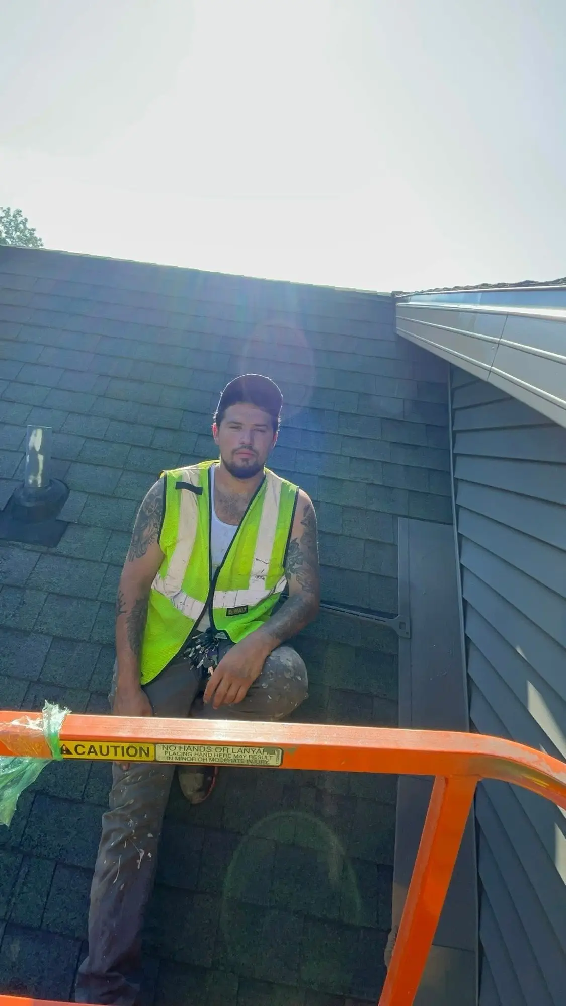 Painter in a hi-vis vest sitting on a shingle roof beside a lift rail, next to gray siding