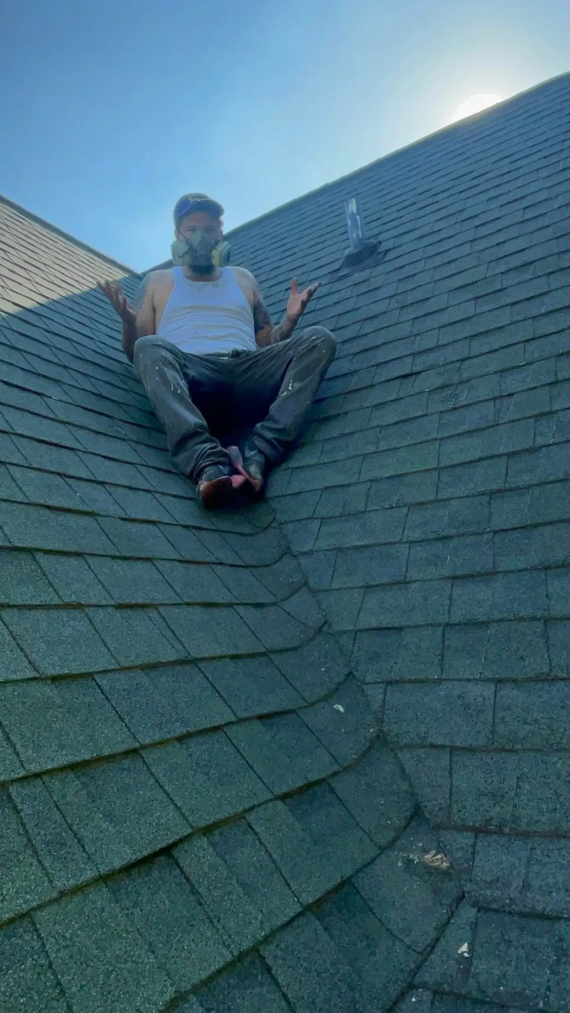 Painter in a respirator sitting on a steep shingle roof against a clear blue sky